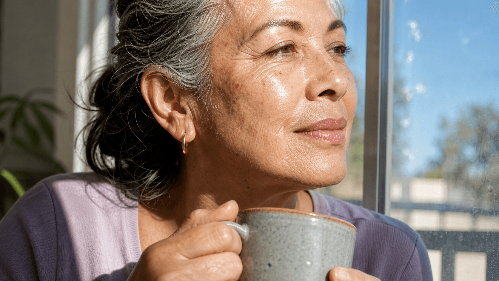 Older adult holding a ceramic mug near a bright window with direct sun on her face.