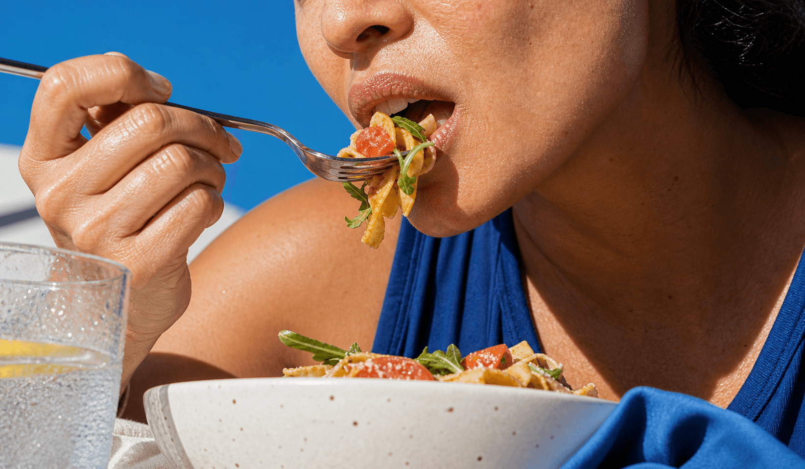 Close crop of an adult eating pasta in hard sunlight with a water glass nearby.